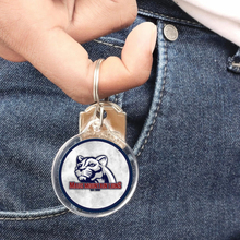 acrylic keychain with a Maus Mountain lions logo held by a finger against denim fabric