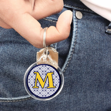 close up of an acrylic photo keychain with a gold initial M and navy blue pattern hanging from a young man's finger