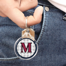 personalized acrylic round keychain featuring a capital letter M in maroon and the Archbishop Curley friar logo in the background hanging from a finger with denim jeans background