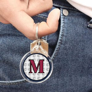 personalized acrylic round keychain featuring a capital letter M in maroon and the Archbishop Curley friar logo in the background hanging from a finger with denim jeans background